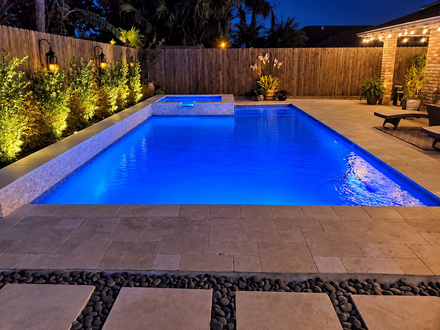 Pool lit blue at night with a raised spa spillover, a tiled raised wall, travertine deck, and lit landscaping